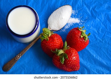 Top Down View Of Three Strawberries With Glass Of White Milk And Teaspoon Of White Sugar On Dark Blue Background