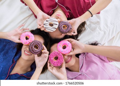 Top Down View Of Three Teenage Girls Having Sleepover Party While Making Funny Face With Donuts And Lying On The Bed At Home