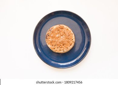 Top Down View Of A Toasted English Muffin With Peanut Butter Spread On A Blue Plate Against A Bright White Background.