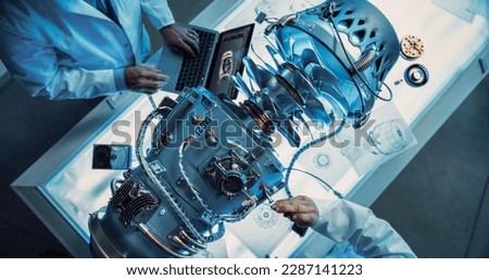 Top Down View of Two Engineers Discussing Work, Talking, Using Laptop Computer and Checking Blueprints in Jet Motor Manufacturing Facility. Scientists Researching Innovative Turbine Engine