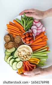 Top Down View Of Two Hands Holding A Large Oval Platter Of Hummus And Crudites.