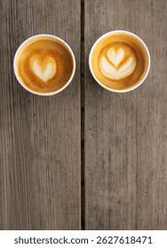 Top down view of two paper coffee cups, placed on a rustic wooden table, with a creamy heart and flower shaped latte art