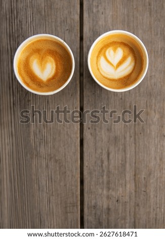 Top down view of two paper coffee cups, placed on a rustic wooden table, with a creamy heart and flower shaped latte art