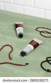 A top down view of two squeeze tubes of cream or paint arranged with decorative beaded jewelry on a soft sage green background with white subway tiles. Tubes have blank white labels for packaging mockup use. Top-view composition with tubes placed diagonally and open space around them. Dark red packaging, white labels and soft green background create a clean color contrast. Beads, cord and small decorative elements visible around the products. Minimal skincare mockup still life with calm bathroom styling. Beauty product photograph focused on packaging, surface texture and self-care objects. Flat lay composition with earthy tones and simple modern arrangement. 
