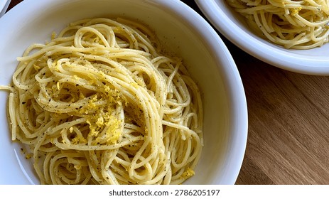 Top down view of two white bowls filled with spaghetti pasta garnished with lemon zest and black pepper on a wooden table.