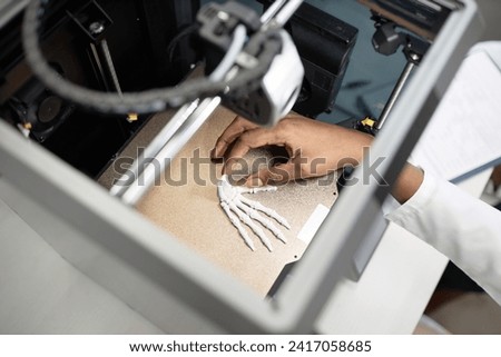 Top down view of unrecognizable technicians hand reaching for illuminated wrist bone model on metal hot bed in enclosed 3D printer