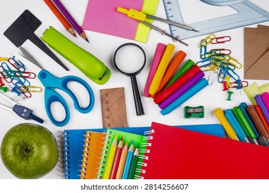 Top down view of various school and office supplies including notebooks, pens, scissors, magnifying glass, and a fresh apple on a white surface.