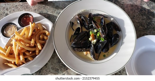 Top Down View Of A White Plate Of French Fries And Mussels On A Picnic Table Under The Sun.