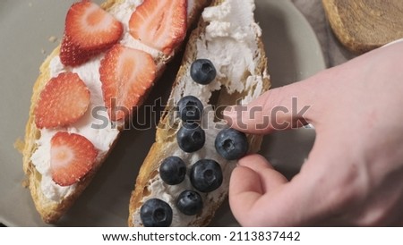 Top down view woman or girl decorate croissants with blueberry on soft cheese. Next to it, there are ready-made pastries with chopped strawberries. Veggie breakfast cooking concept, spicy food.