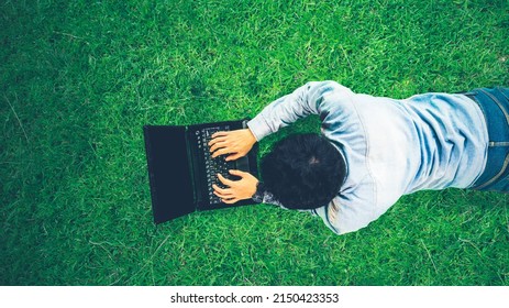 Top Down View Of Young Man Using A Laptop While Lying On The Green Grass At The Park