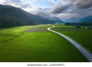 Top drone view of beautiful landscape with alpine mountains, paved highway, hills, fields green grass. Aerial view of beautiful curved rural road in green meadows at dawn in summer in Slovenia