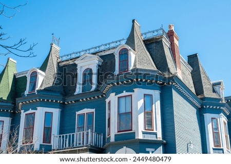 The top exterior corner roof of a historic Victorian building is made of blue clapboard, white and red trim vintage windows, and a large turret in the corner of the decorative heritage building. 