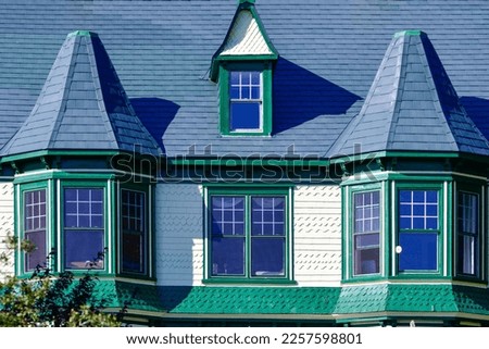 The top exterior floor of a historic Victorian building with two turrets and a peaked dormer window. The glass windows are double-hung, the siding is cedar shakes, and the roof is grey color shingles.
