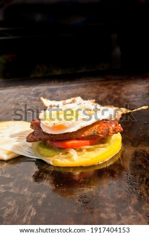 top, front view, medium distance of a Italian sausage, lettuce, tomato, cheese sandwitch cooking on a flat top grill, at a tropical farers market