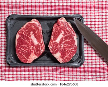 Top Quality Rib Eye Steak On A Black Plastic Tray And Red Table Cloth And Knife. Meat Industry Product. Top Down View.