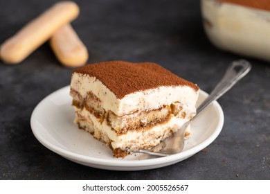 a top shot of a piece of tiramisu with a dish of tiramisu in the back with ladyfingers on a dark background. 