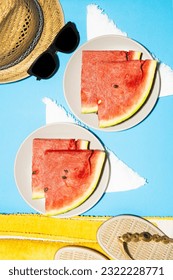 Top vertical view of two plates with slices of watermelon on blue background, straw hat, sandals and beach towel