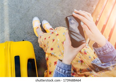 Top Vew Of Rendy Female Traveler With Yellow Suitcase Texting With Smart Device While Sitting On Bus Stop. Sun Glare Effect.