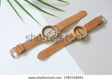 Top view of 2 brown wooden watches on white and brown color background. Green leafs and leather band watch on soft color background.