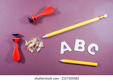 A top view of ABC letters in white, pencil, sharpener residue, yellow color pencil and two sucker darts on a purple background