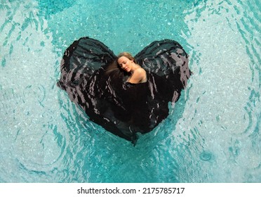 Top View From Above To A Beautiful Sexy Young Woman Floating In Black Cloth Dress Wrapped In Shape Of Valentine Heart In Turquoise Blue Water Of The Swimming Pool, Copy Space