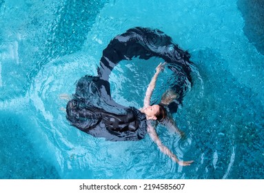 Top View From Above To A Beautiful Sexy Young Woman Floating, Wrapped In Black Cloth Dress In The Form, Shape Of A Ring, Circle In Turquoise Blue Water Of The Swimming Pool, Copy Space