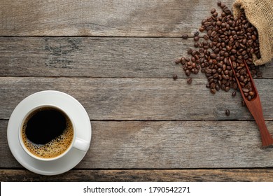 Top View Above Of Hot Fresh Black Coffee With Milk Foam In A White Ceramic Cup With Coffee Beans Roasted In Sack Bag On Wood Rustic Table Background. Flat Lay With Copy Space.