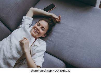 Top view from above young smiling happy woman 20s wear casual white clothes lying on soft grey sofa indoors apartment flat looking camera. Resting on weekends leisure quarantine staying home concept. - Φωτογραφία στοκ