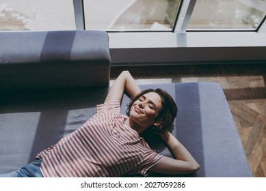Top view from above young woman 20s in casual clothes lying on grey sofa hold hands behind head neck relax take nap near window on weekends rest indoors flat at home People lifestyle leisure concept.