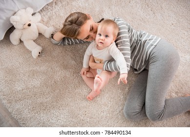 Top-Sicht auf liebevolle Mutter liegt mit kleiner Tochter auf Teppich. Sie umarmt und küsst sie, wie sie saugt – Stockfoto