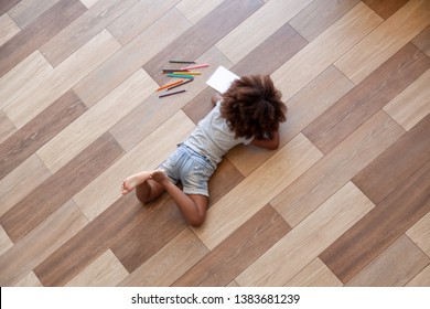 Top View Of African American Small Girl Lying On Home Floor Painting With Colorful Pencils, Black Little Kid Relax Having Fun Coloring Picture In Children Colouring Book, Child Entertainment Indoors