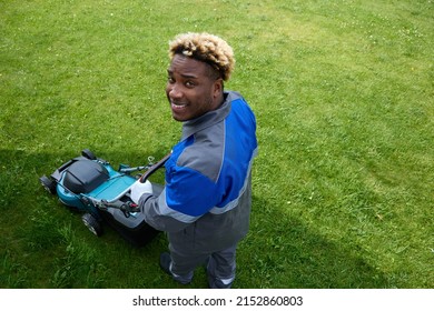 Top View Of An African Man In Overalls Mowing Green Grass In A Modern Garden With A Lawn Mower. A Black Man Looks Up And Smiles Using A Lawnmower In The Backyard. Professional Lawn Care Service.