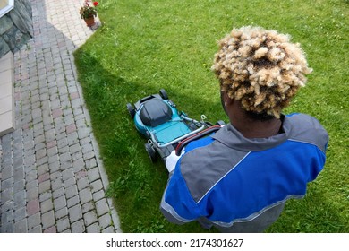 Top View Of An African Man In Overalls Mowing Green Grass In A Modern Garden With A Lawn Mower. A Black Man With An Afro Hairstyle Uses A Lawn Mower In The Backyard. Professional Lawn Care Service.