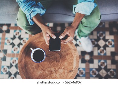 Top view of african man using smartphone while sitting on sofa at home.Concept people working with mobile gadget.Black coffee on the wooden table.Blurred background