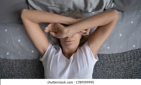 Top View Aged Woman Lying On Bed Woke Up At Night Due Noisy Neighbors. Mature Female Cover Face With Hands Suffers From Insomnia Sleep Disorder, Has Restless Obsessive Thoughts Keep Her Awake Concept