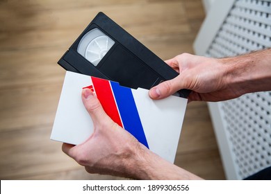 Top View Of A Ale Hand Taking A Videotape, Video Cassette, Vintage VHS Tape Out Of A Case, A Paper Cover, A Box. 