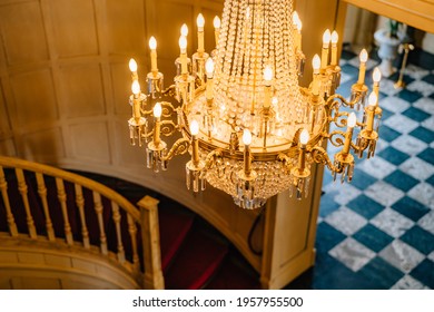 Top view of an antique chandelier. A huge crystal gold chandelier with candles, against the backdrop of a spiral staircase and a checkerboard floor.