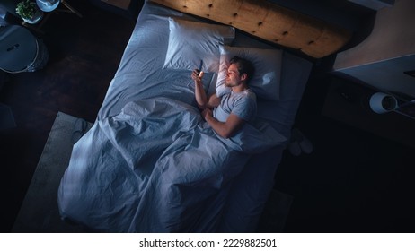 Top View Apartment Bedroom: Handsome Young Man Using Smartphone in Bed at Night. Smiling Guy Browsing Social Media, Dating Apps, Remote Work Software, Doing Internet Online Shopping on Mobile Phone - Powered by Shutterstock