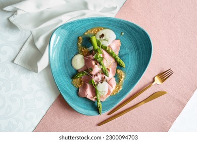 Top view of an appetizer with asparagus, taleggio, peanuts and rosemary ham on a blue plate. Restaurant table with white and pink cloth, white napkin and golden cutlery. - Powered by Shutterstock