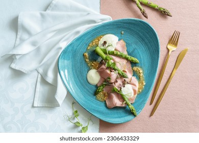 Top view of an appetizer with asparagus, taleggio, peanuts and rosemary ham on a blue plate. Restaurant table with white and pink cloth, white napkin and golden cutlery.  - Powered by Shutterstock