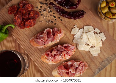 Top view of a appetizer shot on a rustic wooden table, with a varied composition that contains Spanish tapas of Iberico ham, chorizo, slabs of cheese, peppercorns, olives, glass of wine and peppers  - Powered by Shutterstock