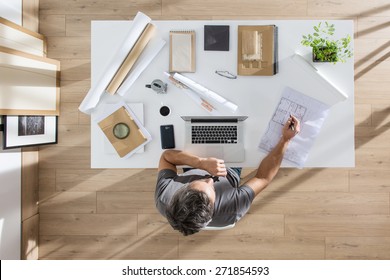 Top View, Architect Sitting At  Desk And Working On His Laptop, There Is Blueprints And Model House On His Table, The Sun Casts Graphics Shadows On The Wood Floor