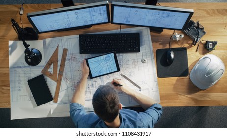 Top View Of Architectural Engineer Draws On His Blueprints, Compares With Tablet Computer With Green Screen, Using Desktop Computer Also. His Desk Is Full Of Useful Objects And Evening Sun.