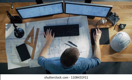 Top View Of Architectural Engineer Working On His Blueprints, Holding Tablet Computer, Using Desktop Computer Also. His Desk Is Full Of Useful Objects And Evening Sun.