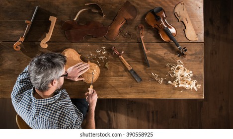 Top view. Artisan luthier working on the creation of a violin. He sits at a wooden table in his workshop, various tools and instrument are placed on the workbench
