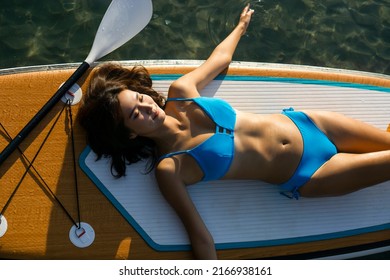 Top view Asian beautiful girl in blue bikini lies on sup board with paddle with closed eyes and rests on water sports board in ocean at sunset background. Woman on Sup top view.