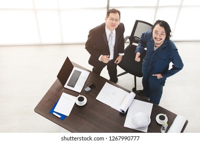 Top View Of Asian Business Man And Male Foreman In Coverall, Happily Look Up At Camera, Satisfy With New Project, On Laptop, Smartphone, Smart Office Background