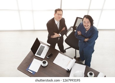 Top View Of Asian Business Man And Male Foreman In Coverall, Happily Look Up At Camera, Satisfy With New Project, On Laptop, Smartphone, Smart Office Background