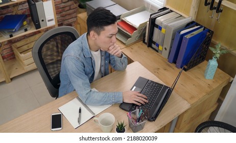 Top View Asian Man Is Writing Down Ideas In His Notebook And Thinking With Hand Under Chin While Typing A Proposal Letter On The Laptop In The Office.