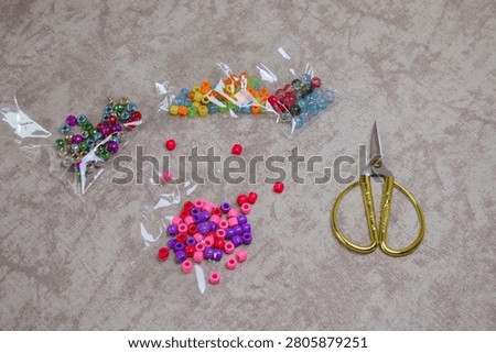 Top view of assorted craft beads in small plastic bags, loose pink and purple beads scattered on the surface, and gold scissors DIY jewelry making hobby, creative pastime, and organized craft supplies